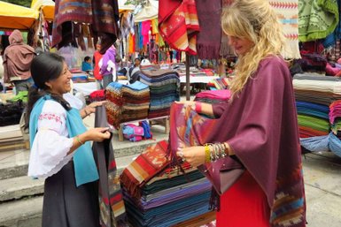 Eine lächelnde Frau in traditioneller Kleidung zeigt einem Touristen bunte Textilien auf einem Markt in Otavalo.