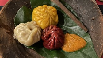 Ein rustikeller Teller mit bunten Momos, umgeben von grünen Blättern und einer kleinen Gabel aus Holz.