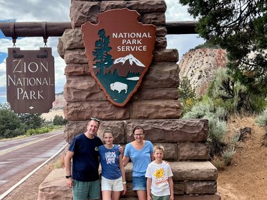 Eine Familie steht lächelnd vor dem Eingang zum Zion Nationalpark, umgeben von beeindruckenden Felsen und Bäumen.
