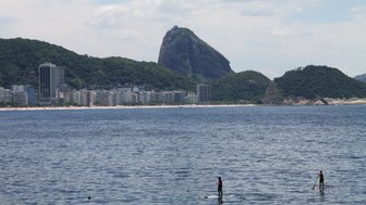 Zwei Paddel-Surfer stehen auf ihren Brettern in ruhigem Wasser, während die Küste von Rio de Janeiro im Hintergrund sichtbar ist.