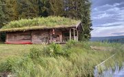 Ein rustikales Holzhaus mit einem begrünten Dach steht inmitten von hohen Bäumen und üppigem Gras am Ufer eines ruhigen Gewässers.