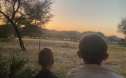 Zwei Kinder stehen nebeneinander und blicken auf eine malerische Landschaft bei Sonnenuntergang, umgeben von Bäumen.