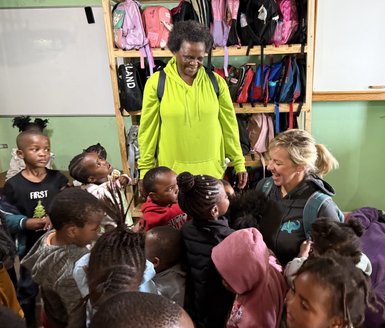 Eine fröhliche Interaktion zwischen einer Lehrerin und einer Gruppe von Kindern in einem Klassenzimmer voller Rucksäcke.