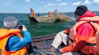 Ein Kind in einer orangefarbenen Schwimmweste fotografiert eine Gruppe von Menschen, die ein Netz auf einem Boot auswerfen.