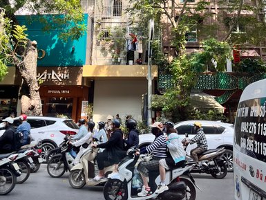 Eine belebte Straße in Hanoi, auf der viele Motorräder und Autos fahren, umgeben von grünen Bäumen und bunten Gebäuden.