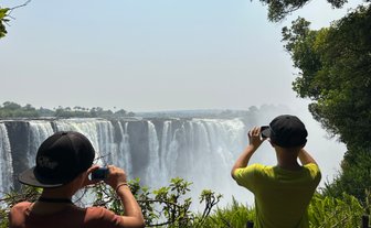 Zwei Kinder stehen am Rand eines Wasserfalls und fotografieren die beeindruckende Aussicht auf die Victoriafälle.