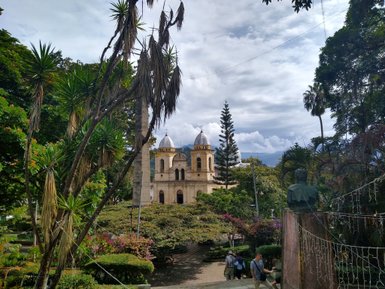 Eine malerische Ansicht eines Parks mit üppigem Grün und einer historischen Kirche im Hintergrund unter einem bewölkten Himmel.