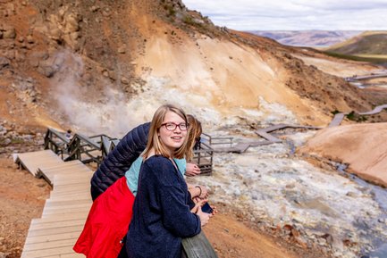 Eine junge Frau mit Brille lächelt, während sie an einem Geländer in einem geothermalen Gebiet steht, umgeben von dampfenden Quellen.
