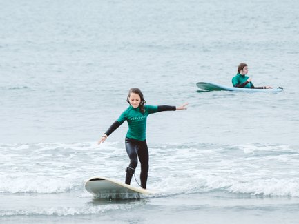 Ein Mädchen surft fröhlich auf einem Surfbrett, während ein anderer Surfer im Hintergrund im Wasser sitzt.