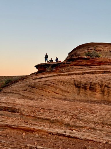 Vier Personen sitzen und stehen auf einer großen, geschwungenen Felsformation, umgeben von einer weiten, offenen Landschaft.