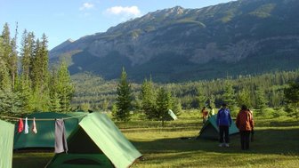 Zwei Personen stehen auf einem Campingplatz mit grünen Zelten, umgeben von hohen Bäumen und majestätischen Bergen im Hintergrund.
