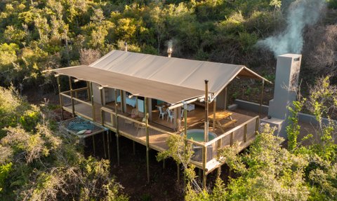 Ein modernes Zelt auf Stelzen, umgeben von üppigem Grün, mit einem rauchenden Kamin und einem einladenden Holzdeck.
