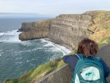 Eine Person mit einem Rucksack steht an der Kante der Klippen und blickt auf die raue See und die zerklüfteten Felsen.