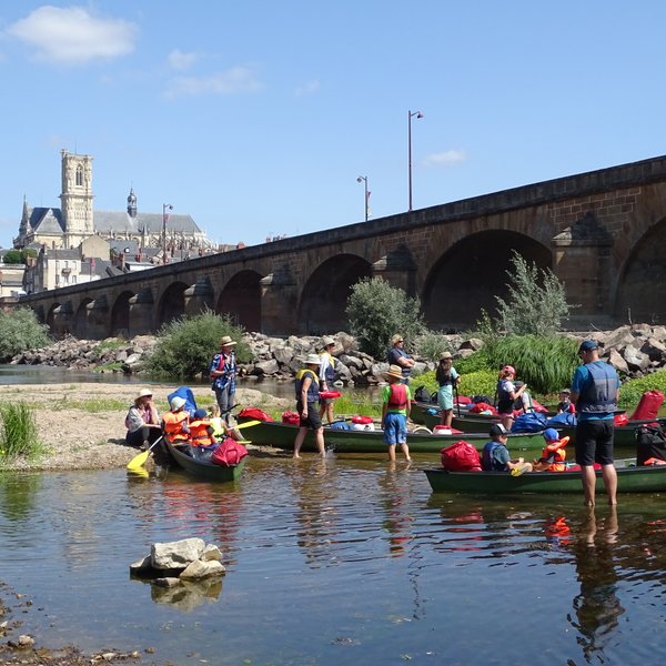 Eine Gruppe von Menschen in Kanus bereitet sich auf eine Kanutour an einem sonnigen Flussufer vor, mit einer Brücke im Hintergrund.