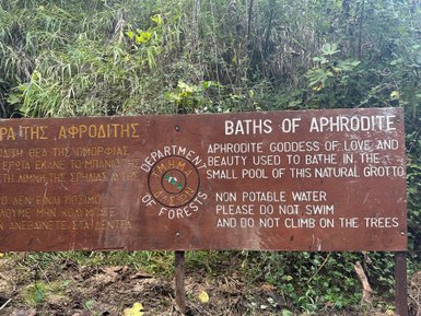 Ein Holzschild beschreibt die Bäder der Aphrodite, umgeben von üppigem Grün und einem kleinen natürlichen Pool.