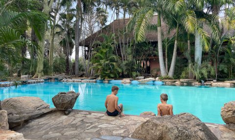 Zwei Jungen sitzen am Rand eines blauen Pools, umgeben von üppigen Palmen und Felsen, während sie die Aussicht genießen.