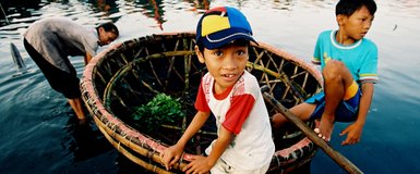 Ein Junge mit einer bunten Mütze sitzt in einem Korbboot, während ein anderer Junge nach etwas im Wasser schaut.
