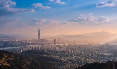 Die Skyline von Seoul bei Sonnenuntergang, mit dem markanten Lotte World Tower, umgeben von sanften Hügeln und Wolken.