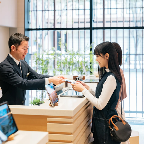 Ein freundlicher Hotelmitarbeiter überreicht einer jungen Frau an der Rezeption eine Karte, während sie lächelt.
