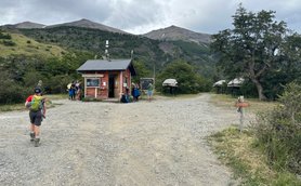 Ein Wanderer mit Rucksack geht auf einem Schotterweg zu einer kleinen roten Hütte in der Natur Patagoniens.