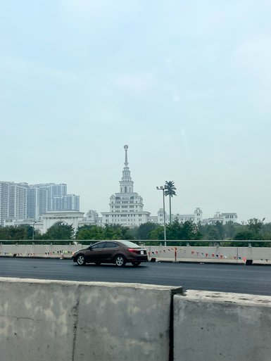 Ein braunes Auto fährt auf einer Straße, während im Hintergrund ein markantes Gebäude mit einem hohen Turm sichtbar ist.