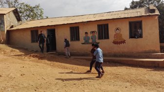 Eine Gruppe von Kindern spielt fröhlich auf einem staubigen Weg vor einem einfachen Schulgebäude mit bunten Wandmalereien.
