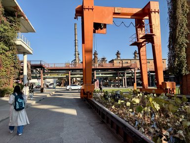 Eine Person in einem langen, weißen Mantel geht auf einem gepflasterten Weg, umgeben von alten Industriegebäuden und Pflanzen.