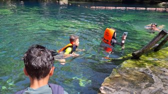 Ein Junge mit einer orangefarbenen Schwimmweste steht am Ufer eines klaren Cenotes, während andere im Wasser schwimmen.