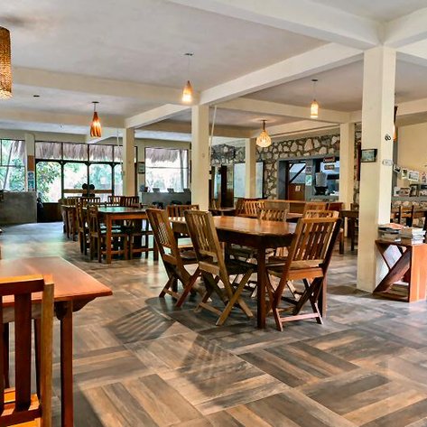 Ein einladendes Restaurant mit Holztischen und Stühlen, umgeben von großen Fenstern, die viel Tageslicht hereinlassen.