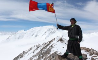 Ein Mann in traditioneller mongolischer Kleidung steht auf einem Felsen und hält die mongolische Flagge hoch in den Bergen.