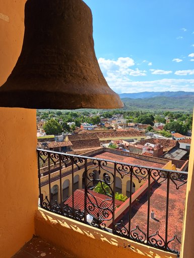 Eine große Glocke hängt über einem Balkon mit Blick auf eine malerische Stadtlandschaft und grüne Hügel im Hintergrund.
