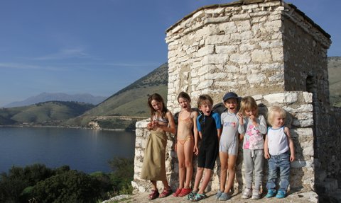 Eine Gruppe von fröhlichen Kindern steht an einem steinernen Gebäude am Wasser, umgeben von grünen Hügeln und blauen Himmel.