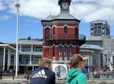 Zwei Jungen stehen am Wasser und schauen auf ein historisches Gebäude mit einer Uhr und einem roten Anstrich.