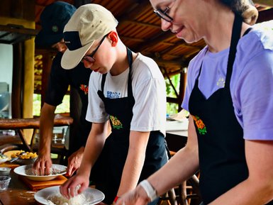 Drei Personen in Kochschürzen arbeiten voll konzentriert am Tisch mit Teig, während sie entspannt miteinander kommunizieren.