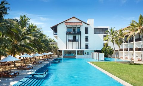 Ein modernes Hotelgebäude mit einem großen Pool, umgeben von Palmen und Liegen, strahlt tropische Entspannung aus.