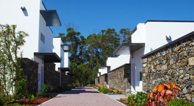 Eine sonnige Straße zwischen modernen, weißen Ferienhäusern, umgeben von üppigem Grün und bunten Pflanzen.