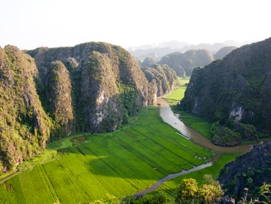 Eine atemberaubende Landschaft mit hohen, bewaldeten Felsen, die sich über saftige grüne Reisfelder und einen sanft fließenden Fluss erstrecken.