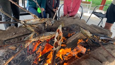 Über dem offenen Feuer werden mehrere Fische auf Stöcken gegrillt, während Menschen gespannt zuschauen.
