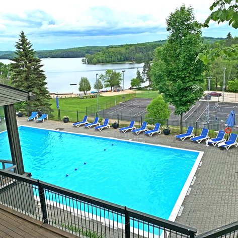 Ein klarer Pool mit blauen Liegen umgeben von Bäumen, Blick auf einen ruhigen See und Sportplätze im Hintergrund.