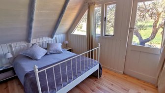 Ein gemütliches Schlafzimmer mit einem weißen Bett, gestreiften Kissen und großen Fenstern, die auf einen grünen Garten blicken.