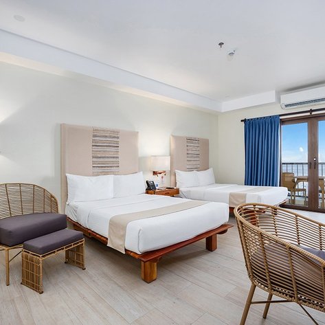 Ein helles Hotelzimmer mit zwei Betten, einem Schreibtisch und einem gemütlichen Stuhl, der Blick auf das Meer ist sichtbar.