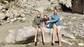 Drei Kinder und eine Frau sitzen auf einem großen Stein am Ufer eines schmalen Flusses, umgeben von steilen Felsen.