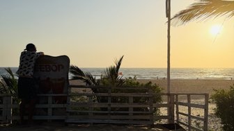 Ein Mann lehnt an einem Schild und blickt auf den Sonnenuntergang über dem Meer, umgeben von Palmen und Sand.