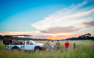 Eine Gruppe von fünf Personen steht in einem weiten Grasfeld, während die Sonne am Horizont untergeht.