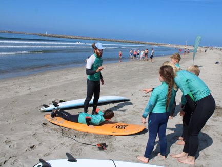 Eine Gruppe von Kindern in Neoprenanzügen beobachtet einen Surflehrer, der Anweisungen am Strand gibt.