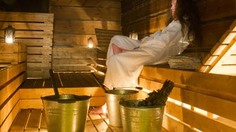 In einer warmen Sauna sitzt eine Person in einem weißen Bademantel entspannt auf einer Holzbank, umgeben von Eimern.