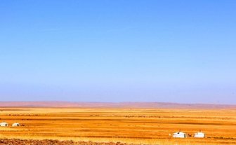 Eine weite, goldene Steppe erstreckt sich unter einem klaren blauen Himmel, mit wenigen Jurten in der Ferne.