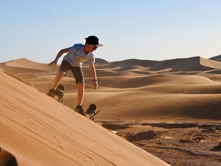 Kind gleitet mit einem Sandboard die sandigen Dünen der Wüste hinunter – Familienreise nach Marokko