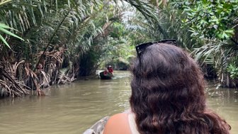 Eine Person mit lockigem Haar sitzt in einem Boot und genießt die ruhige Fahrt durch einen grünen Wasserweg im Mekong-Delta.