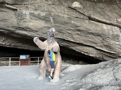 Ein Junge umarmt eine große Statue eines prähistorischen Tieres in einer Höhle, umgeben von grauem Kies und Felsen.